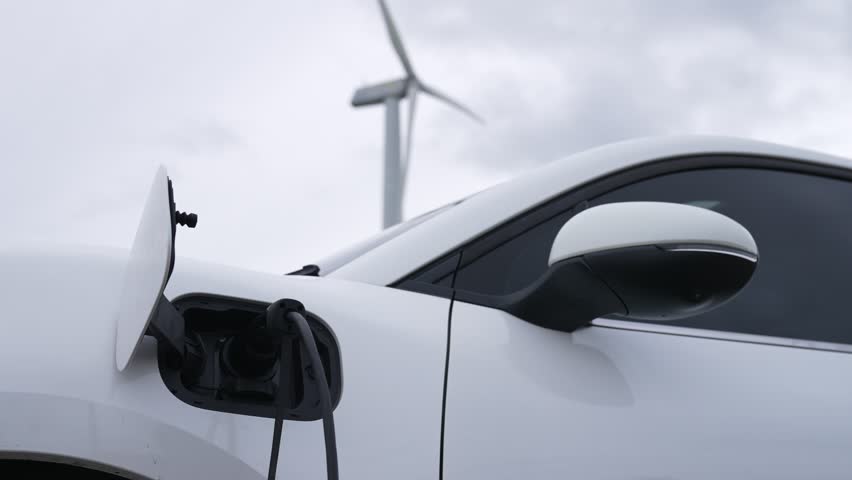 Progressive future energy infrastructure concept of electric vehicle being charged at charging station powered by green and renewable energy from a wind turbine in order to preserve the environment. | Shutterstock HD Video #1099878445