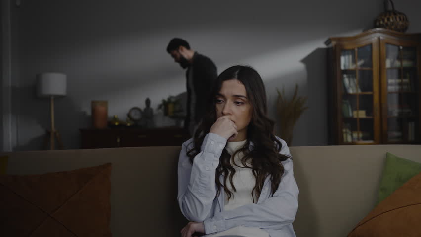 Hopeless young Caucasian woman sitting on the sofa at home, while her husband screaming at her, expressing aggression. Domestic violence and emotional abuse concept. People and social issue. Royalty-Free Stock Footage #1099880491
