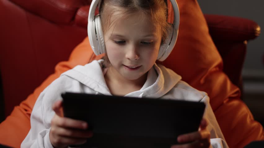 Excited teen girl plays game at home on digital tablet technology device sitting on sofa. Emotional child in headphones holds pad computer surfing internet. Children tech addiction concept | Shutterstock HD Video #1099881549