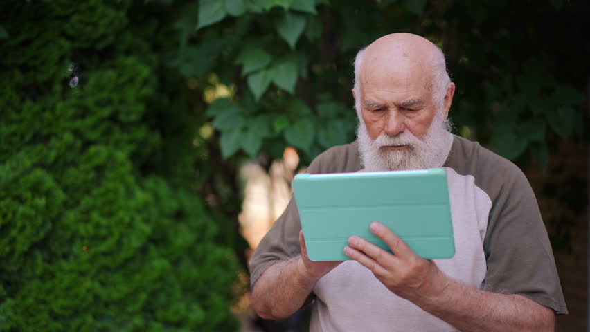 Portrait of concentrated serious senior man surfing internet on digital tablet sitting in summer spring garden on the right. Focused Caucasian male retiree messaging online outdoors thinking