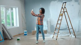 Young african american woman is making video call using her mobile phone. Black female in checkered shirt communicates remotely through camera and shows interior of the room during renovation.