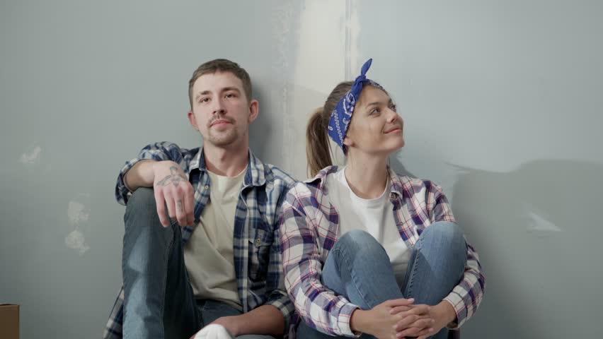 Tired but happy young man and woman are sitting on floor near gray wall and admiring renovation in apartment. A loving couple is embracing and lovingly admiring the results of their work. Royalty-Free Stock Footage #1099886031