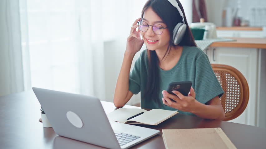 Relax young asian woman wear wireless headphones having fun listen music in smartphone and laptop, Female enjoys listening chill music audio sound feeling no stress at home. | Shutterstock HD Video #1099892285