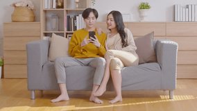 Asian young couple lover sit on couch or sofa looking short video clips in social media having fun and laughing together. Happiness family spending time with good moment together in holiday at home.