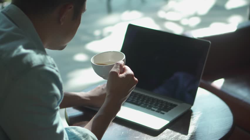 A young businessman using laptop while sitting in cafe. Attractive young man with cup of coffee texting on laptop. | Shutterstock HD Video #1099895989