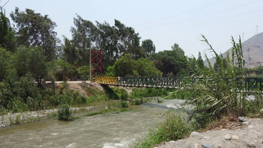 Drone flies towards a bridge over a river. Behind the bridge are many tall trees. The bridge is colored red, yellow and green. Located in Cineguilla in Lima, Peru.