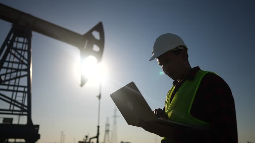 Oil business. a worker works next to lifestyle an oil pump holding a laptop. industry business oil and gas concept. engineer studying the level of oil production on a laptop silhouette at sunset