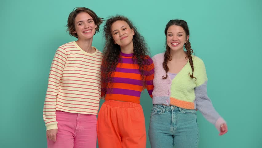 Three smiling young girls hugging and waving hands at the camera in the turquoise studio