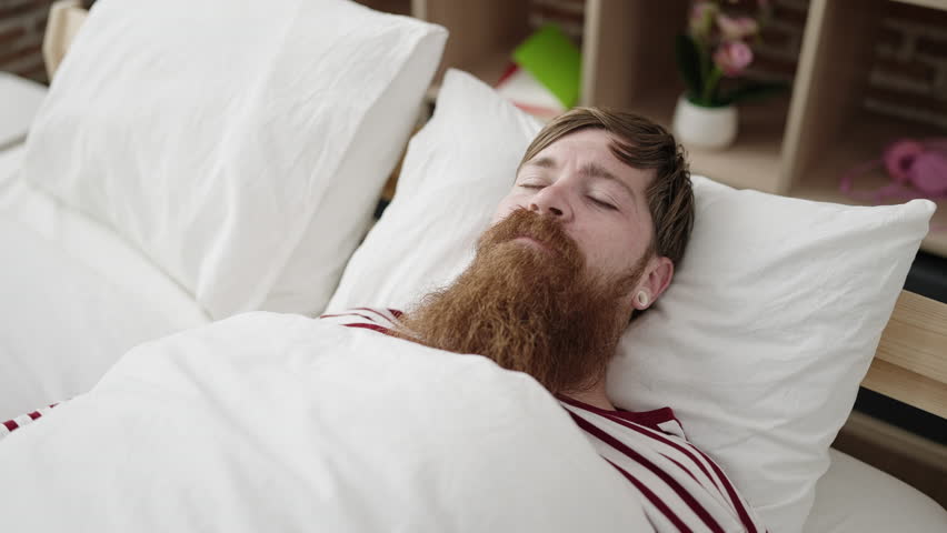 Young redhead man somnambulist sitting on bed at bedroom