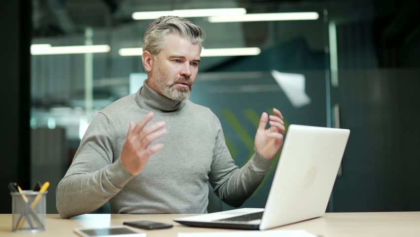 Confused mature gray haired bearded businessman is solving a complex problem while working on a laptop while sitting at a desk in modern office. Disappointed entrepreneur can't find the right solution | Shutterstock HD Video #1099915297