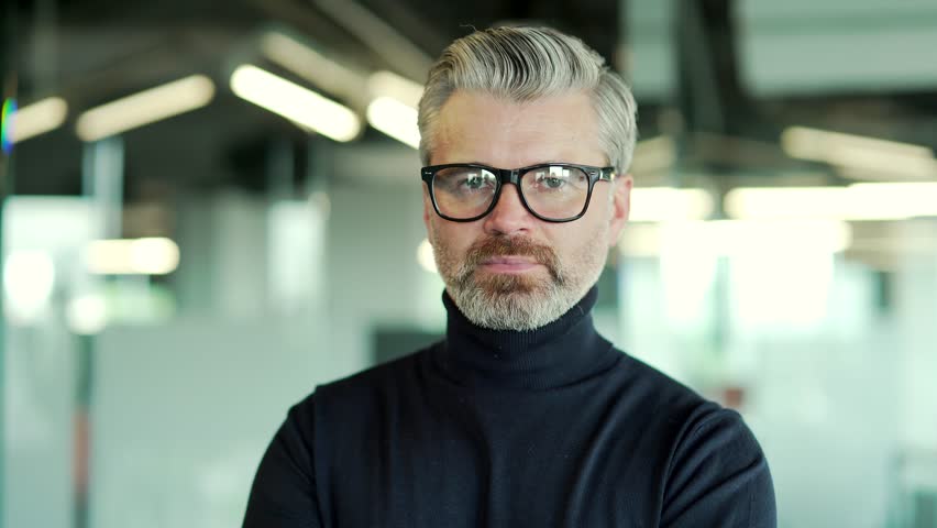 Close up portrait of serious mature gray haired bearded man in glasses looking at camera while standing in modern office. Middle aged confident businessman with a concentrated look and crossed arms | Shutterstock HD Video #1099915303