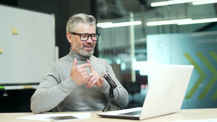 Smiling mature gray haired bearded man is having a video call using a laptop while sitting at a desk at a workplace in a modern office. Happy employee happily shares his ideas at an online conference | Shutterstock HD Video #1099915327