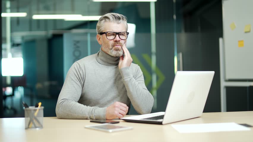Mature gray haired bearded businessman wearing glasses has a toothache while sitting at a desk at a workplace in modern office. Tired middle aged entrepreneur holding hand on cheek massaging sore spot | Shutterstock HD Video #1099915333