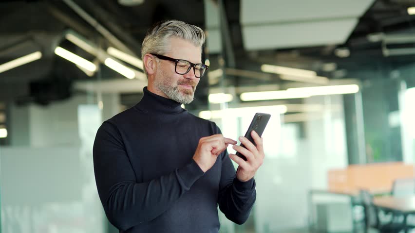 Excited happy mature gray haired bearded man wearing glasses read good news on his smartphone while standing in modern office. Businessman celebrates success by holding his hand in a triumphal gesture Royalty-Free Stock Footage #1099915341