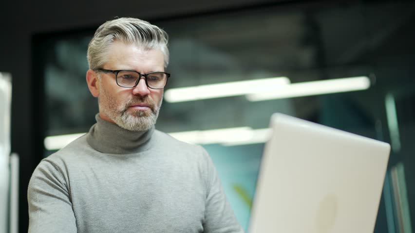 Close up of concentrated mature gray-haired bearded businessman in glasses typing on laptop at workplace in modern office. A confident entrepreneur seriously thinks about what to write to a client | Shutterstock HD Video #1099915349