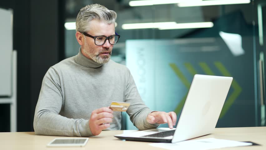 Sad mature gray haired bearded businessman having problem typing credit card number on laptop at workplace in office. Entrepreneur is angry because he faced fraud, money was stolen from his account | Shutterstock HD Video #1099915351
