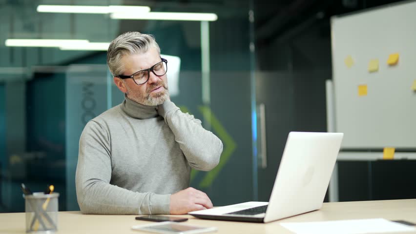 Mature gray haired bearded businessman wearing glasses has neck pain while sitting at desk at workplace in modern office. A tired middle aged entrepreneur rubs and massages his muscles with his hands | Shutterstock HD Video #1099915357