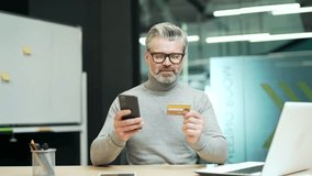 Smiling mature gray haired bearded businessman wearing glasses is typing credit card number on smartphone while sitting at workplace in modern office Happy confident entrepreneur doing shopping online - Powered by Shutterstock - Get 15% off with code: PIKWIZARD15