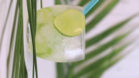 Lemonade with lime, mineral water and crushed ice. Pouring lemonade into a glass from a carafe. Summer composition with refreshing drink and palm leaves