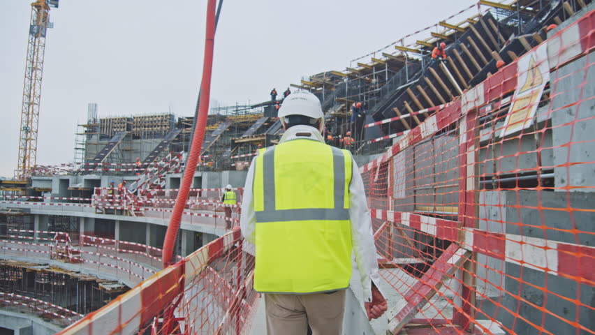 Contractor with color safety vest walks along tribune of sports stadium with red white fences at construction site backside view | Shutterstock HD Video #1099917589