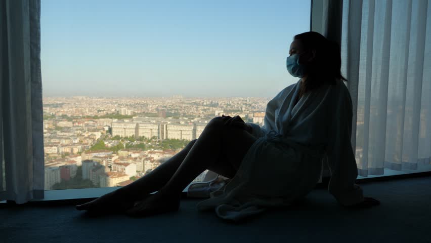 Quarantine isolation upon arrival in another country. A woman sits on the floor by the window and looks sadly at city. She is wearing a bathrobe and medical mask on her face. Strict rules for covid-19 Royalty-Free Stock Footage #1099930251