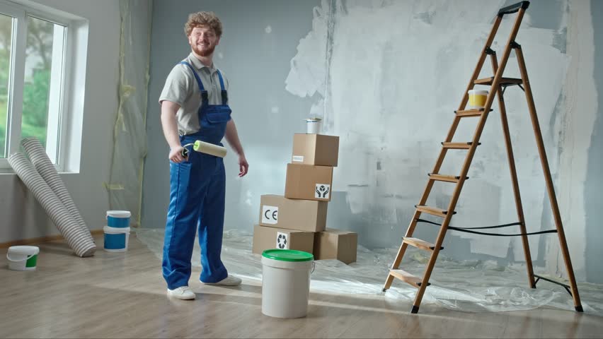 Foreman or painter in blue construction overalls and white helmet with bucket of paint in his hands. Redhead man taking paint roller and planning to paint walls in renovated apartment. | Shutterstock HD Video #1099936567