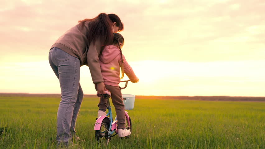 cheerful mother teaches little child ride bicycle sunset. happy family concept. mom with child girl daughter rides bike. girl pedals bicycle. kid rides green grass. spin wheel two-wheeled bicycle