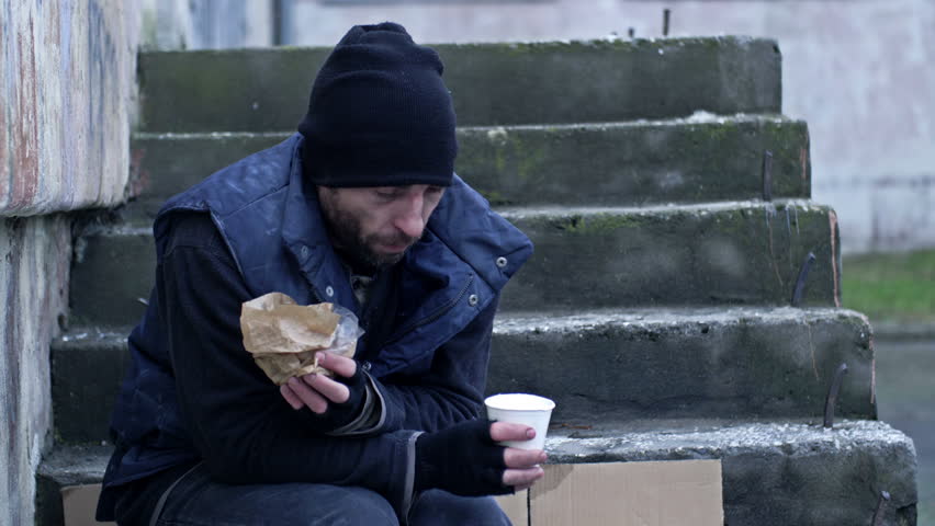 Dirty, cold and poorly dressed man sits on the steps outside and eats something.