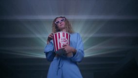 Cheerful caucasian woman dancing, eating popcorn, watching music videos on a projector at home. Low-angle shot of a happy blonde woman wearing glasses having fun. High quality 4k footage