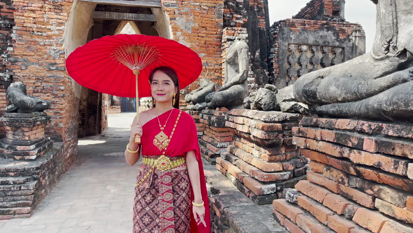 young woman wearing traditional red Thai dress and golden accessories stands holding a traditional umbrella in the historical site Wat Chaiwatthanaram temple. Thai national costume