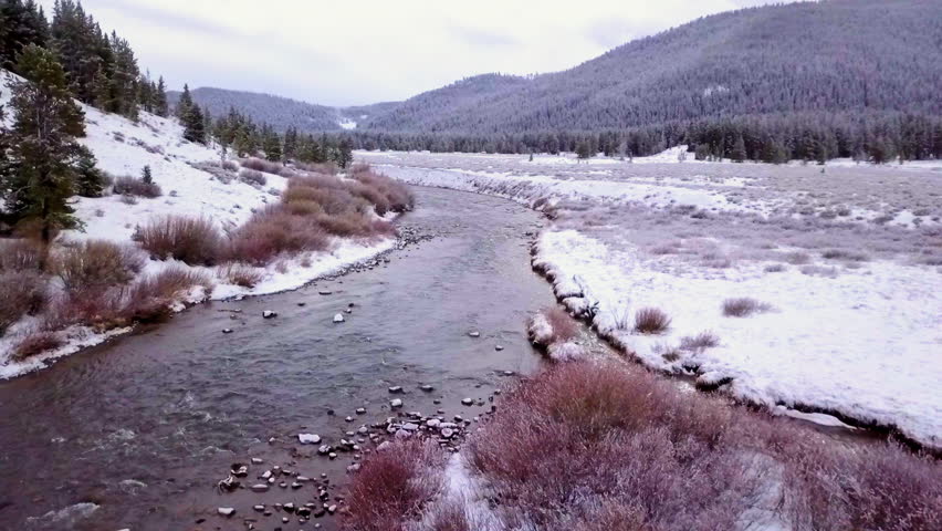 Aerial: Drone Backward Shot Of River Flowing By Snow Covered Mountain Against Sky - West Yellowstone, Montana