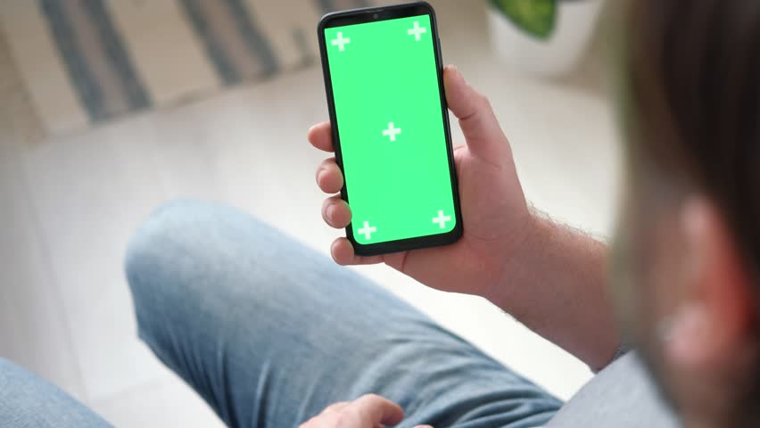 Young man sitting at home holding smartphone green mock-up screen in hand. Male person using chroma key mobile phone. Vertical mode. Touching, swiping display, tapping, surfing Internet social media | Shutterstock HD Video #1099955289
