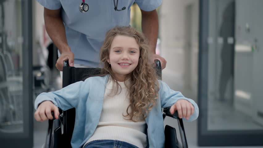 Young caregiver pushing little girl on wheelchair at hospital corridor, having fun together. Royalty-Free Stock Footage #1099957321
