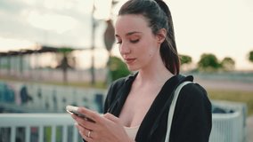 Young woman with freckles and long ponytail wearing black hoodie looking through social networks, photo, video on smartphone, raises her head and smiles