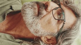 VERTICAL VIDEO: Clouse-up, middle-aged man with gray hair and beard wearing casual clothes sits on bench. Mature gentleman in eyeglasses turns his head and looks into the camera and smiles