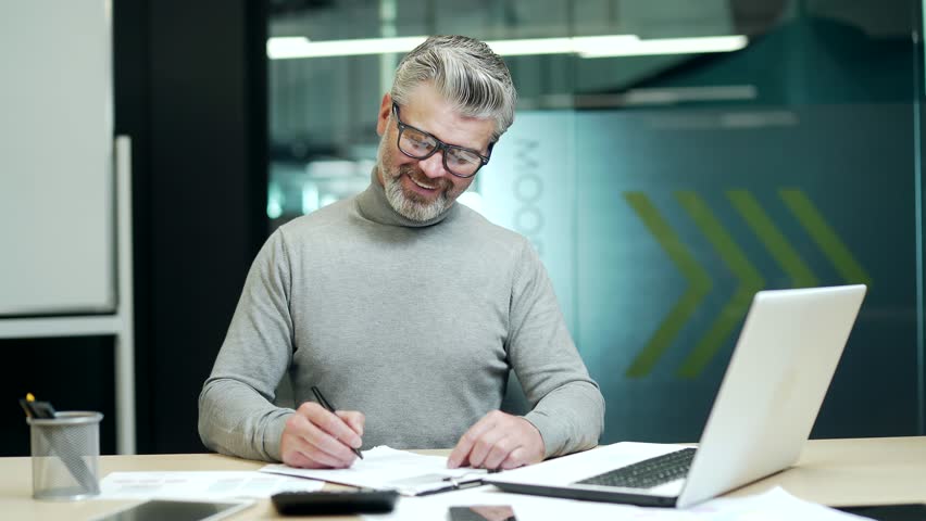 Smiling mature gray haired bearded businessman in glasses making notes in notebook at desk at workplace in modern office. Happy entrepreneur owner making plans for the day showing hand gesture yes | Shutterstock HD Video #1099963141