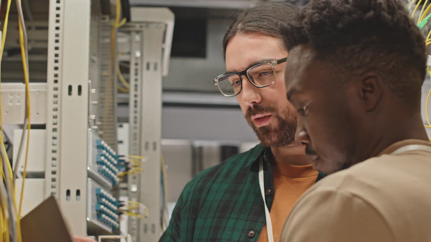 Two diverse maintenance workers using laptop while controlling work of supercomputer in rack server cabinet of data center Royalty-Free Stock Footage #1099967509