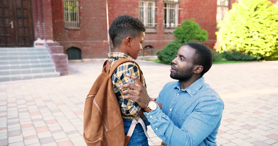 Portrait of African American handsome young man talking with cute son at schoolyard outside. Nice male junior student with school bag speaking with father near school. Family concept