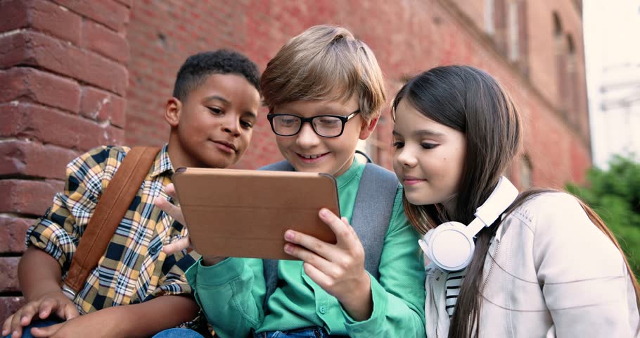 Close up portrait of mixed-races pupils playing on tablet outdoor near school. Happy Caucasian boy and girl tapping and scrolling on device. African American kid talking at schoolyard. Friends concept | Shutterstock HD Video #1099967837