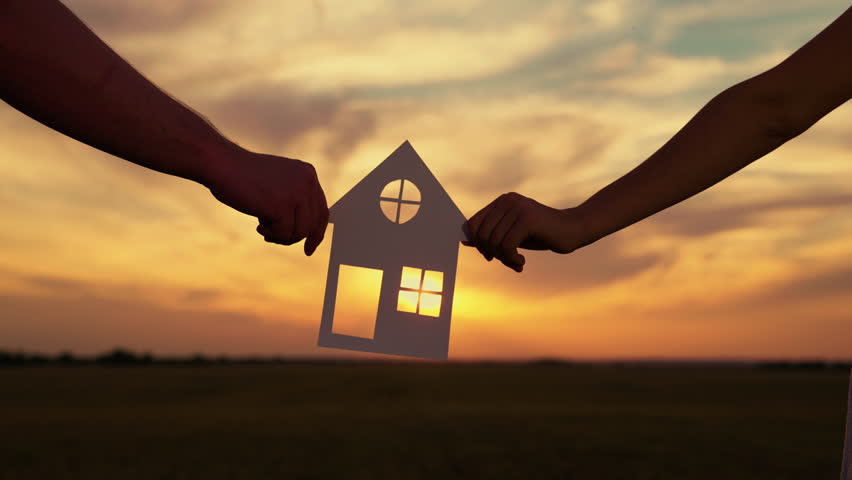 Familys hands are holding paper house at sunset, sun is shining through window. Symbol of house, happiness. Concept of building house for family. Dream to buy house. Home for children and parents Royalty-Free Stock Footage #1099973513