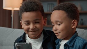 Two happy little African American siblings boys children kids little with mobile phone smartphone talk video call virtual app conference laugh playing game online talking laughing sit at couch at home
