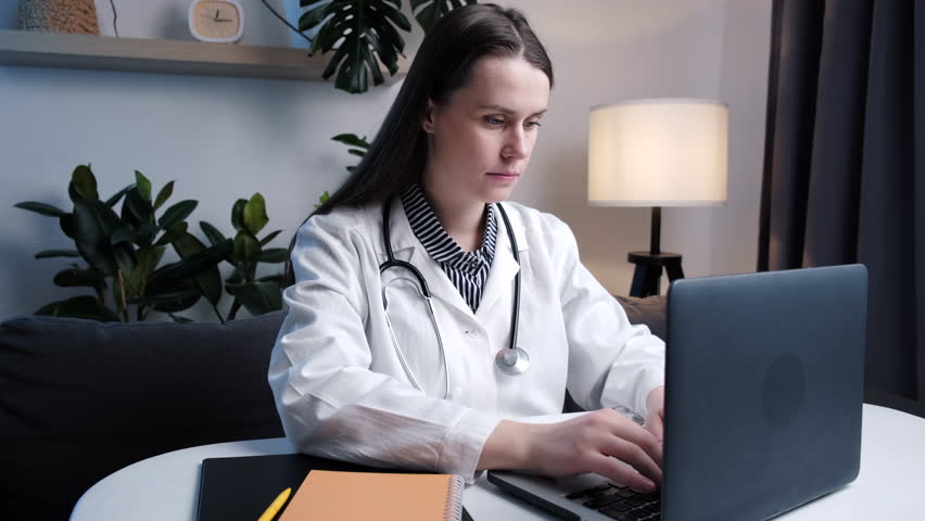 Focused young woman doctor physician wearing white coat and stethoscope using pc laptop computer sitting at desk. Female professional medical consultants patient distantly in online chat at workplace | Shutterstock HD Video #1099978273