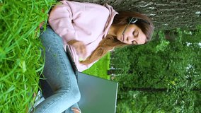 Young woman with laptop and headset having online conversation while sitting in city park - watching educational webinar on laptop. Slow motion vertical footage.