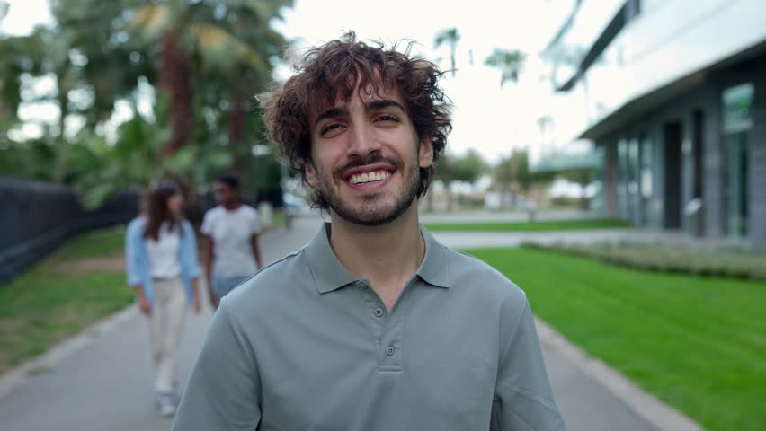 Front view of confident smiling young adult man looking at camera walking on the street with friends on the background in summer. Millennial people and youth concept