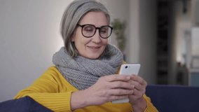 Smiling mature woman watching videos on smartphone sitting on cozy sofa, subscribing to watch new videos without advertisement and additional payments, possibility to download and watching offline