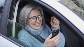 Trendy mature woman in stylish eyeglasses and with fashionable hairdo laughing watching funny videos online on smartphone, lady dancing, trying to repeat social network trend
