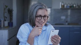Active mature lady in stylish eyeglasses looking performance of favourite music band in internet on smartphone and dancing, having fun and enjoying, good mood
