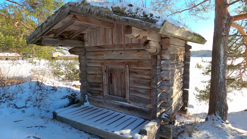 Old small wooden log cabin from 1800s standing on a shoreline of a lake in a bright winter day in Finland