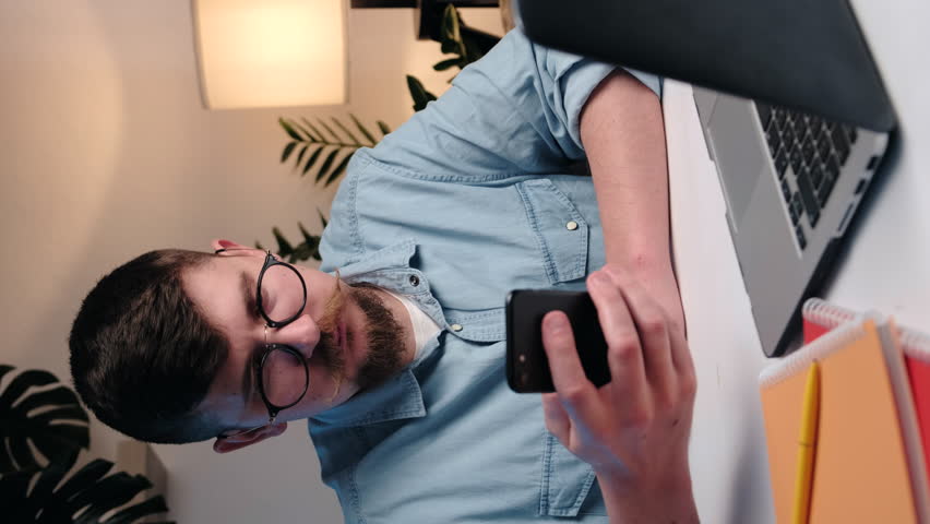 Vertical video of serious young man using laptop and modern smartphone in office. Smart executive multitasking checking synced apps at work. Mobile and pc technology data synchronization for business. Royalty-Free Stock Footage #1099986949