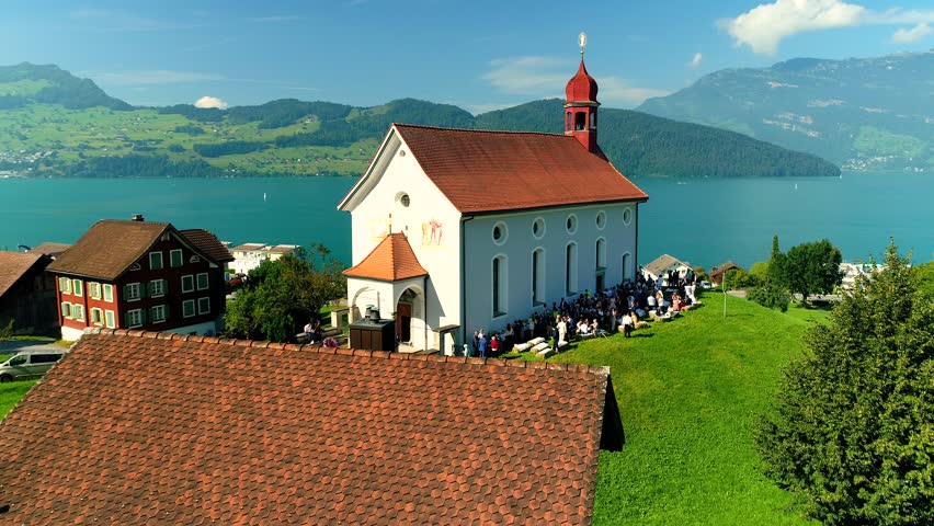 A drone shot of crowd next to chapel with a lake and mountains in the background on a sunny day